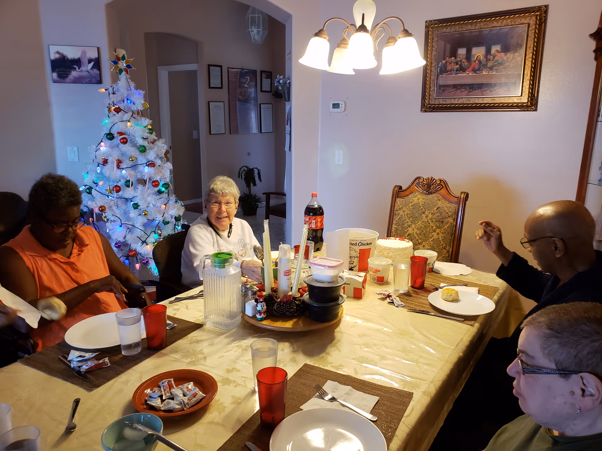 Four elderly people sitting around a dining table with plates, cups, and food items including a cake and a bucket of fried chicken. A decorated white Christmas tree with colorful lights is visible in the background. The room has a chandelier light fixture and a framed picture on the wall.