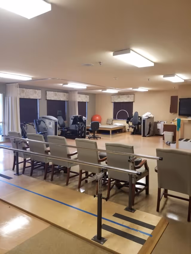A rehabilitation room with parallel bars for walking exercises, several chairs lined up, exercise equipment, a therapy bed, and a large red exercise ball. The room has beige walls, multiple windows with blinds and valances, and bright overhead lighting.