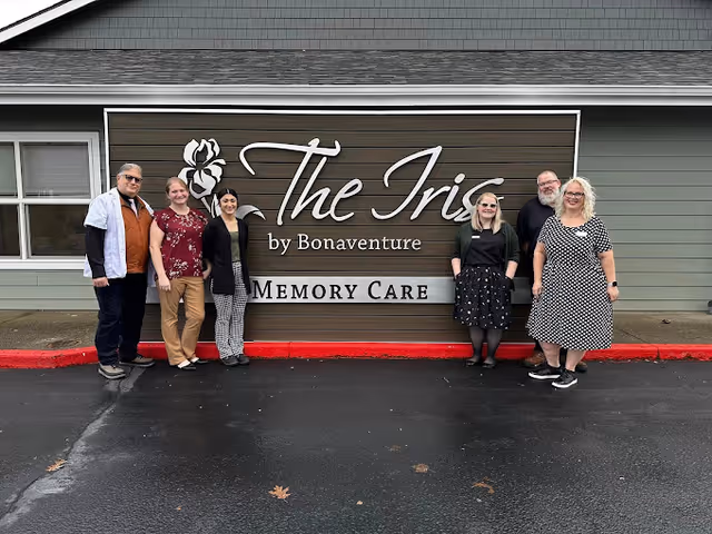 Six people standing in front of a large sign that reads 'The Iris by Bonaventure MEMORY CARE' mounted on the exterior wall of a building with green siding and a window to the left.