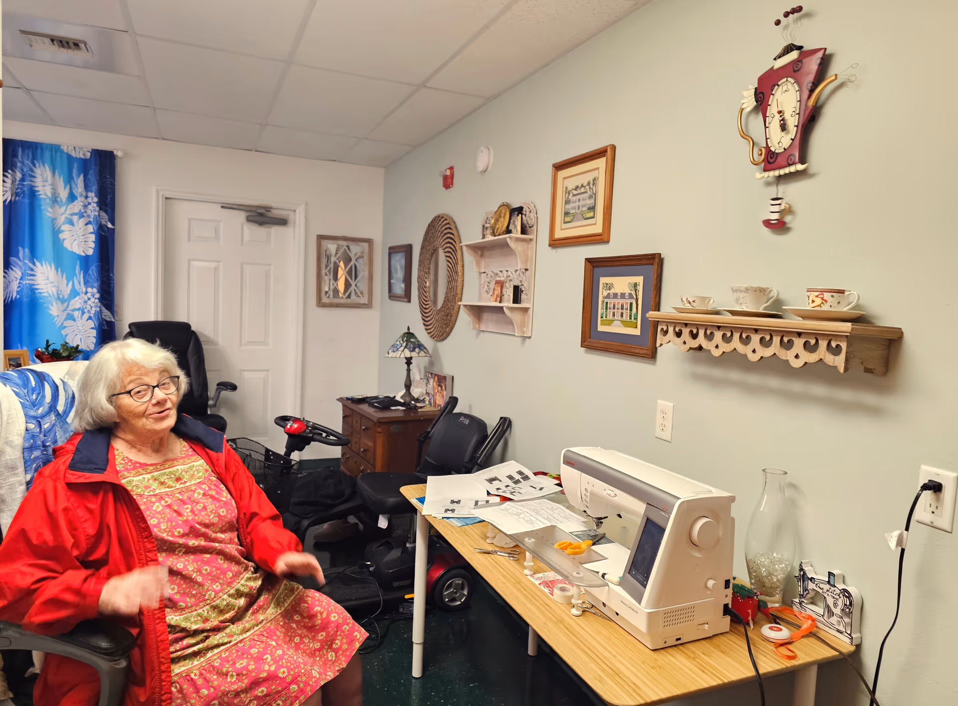 An elderly woman wearing glasses, a red jacket, and a floral dress sits in a chair in a cozy room. The room has a sewing machine on a wooden table with scissors, papers, and sewing supplies. The wall behind the table is decorated with framed pictures, a round woven mirror, a small shelf holding teacups, and a whimsical clock. A mobility scooter and a small wooden dresser with a lamp are also visible in the background.