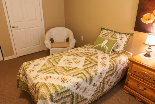 A small bedroom with a single bed covered in a green and white patterned quilt. There is a green pillow with a bird design on the bed. Next to the bed is a wooden nightstand with a lamp and a floral painting on the wall above it. A white wicker chair with a beige cushion is placed near a closed white door.