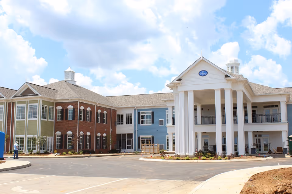 Front exterior of a two-story senior living facility with tall white columns, mixed brick and clapboard siding, and a circular drive under a partly cloudy sky.
