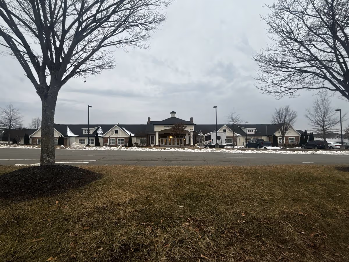 Wide frontal view of a single-story senior living facility with a central entrance, parked cars, and snow along the curb under a gray sky.