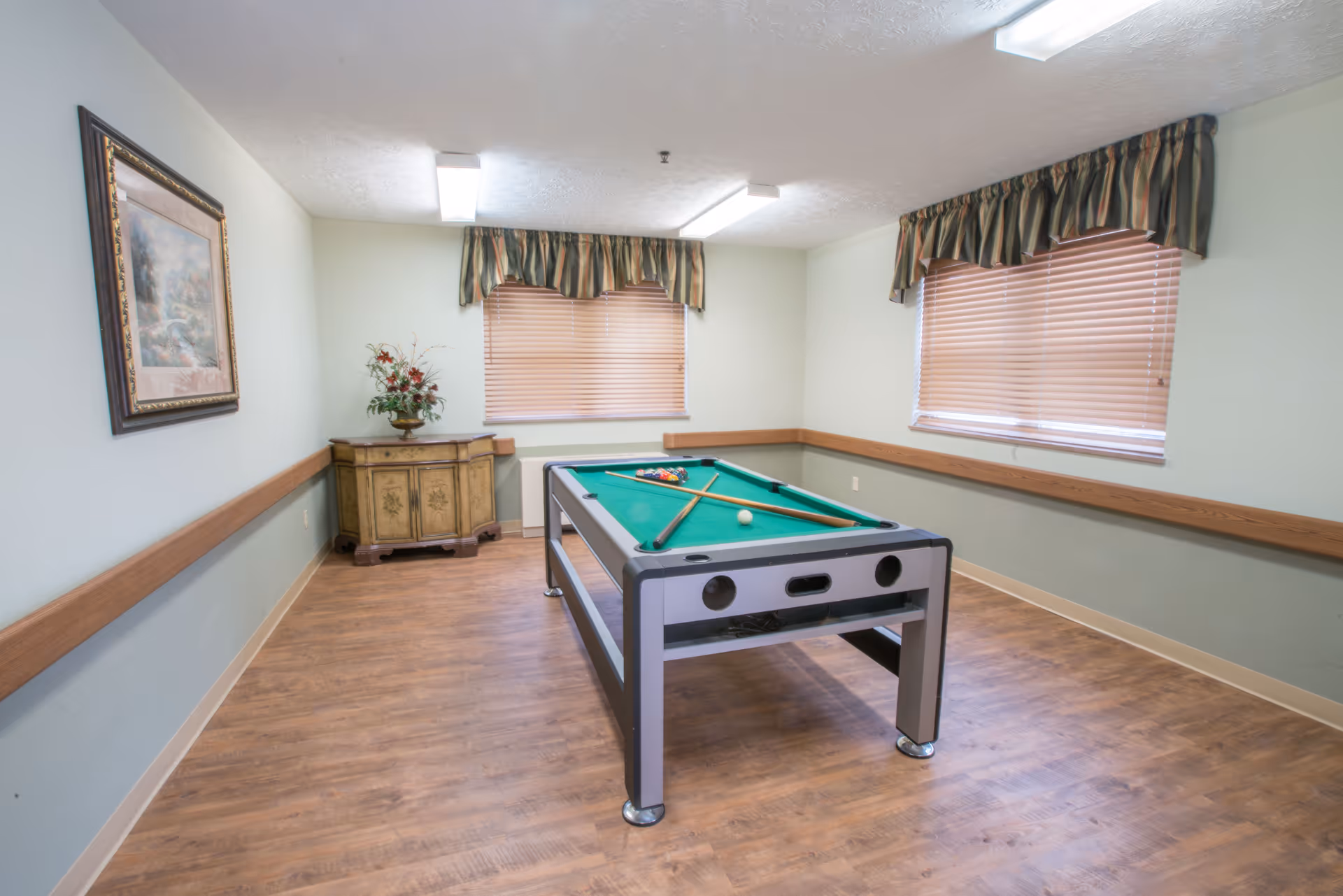 A room with a pool table in the center, two pool cues and balls arranged on the table. The room has wooden flooring, light green walls with wooden trim, two windows with blinds and valances, a framed painting on the left wall, and a small decorative cabinet with a flower arrangement in the corner.