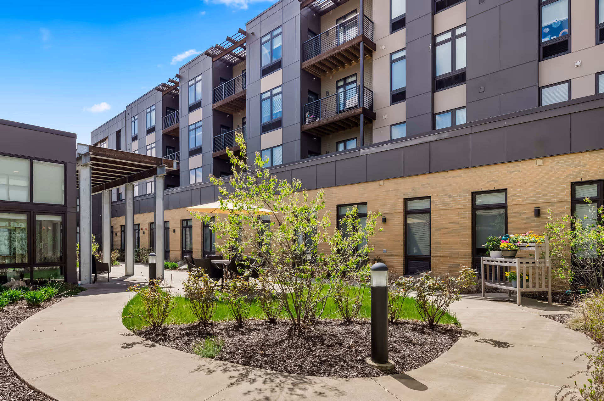 Outdoor courtyard area of a senior living facility with a curved concrete walkway, landscaped garden beds with bushes and small trees, patio tables with umbrellas, and a multi-story building with balconies in the background under a clear blue sky.