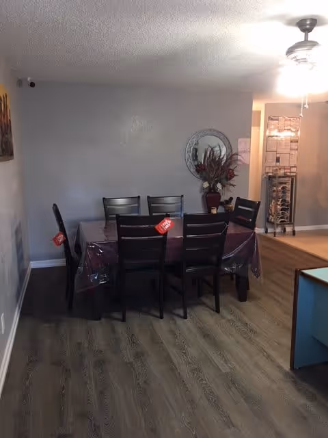 A dining area with a rectangular table covered by a plastic tablecloth and six dark wooden chairs around it. A decorative round mirror and a vase with dried flowers are on the wall behind the table. The floor is wood laminate, and a ceiling fan with a light is visible above. There is a metal rack with items in the background near a doorway.