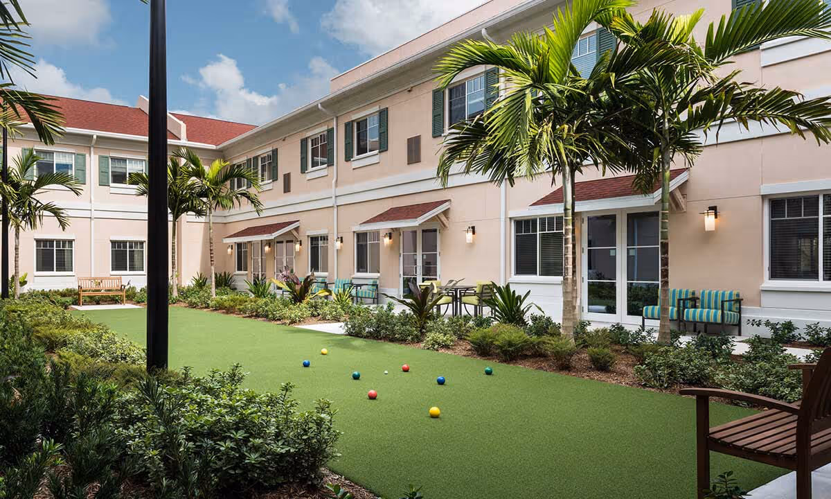 Outdoor courtyard area at Alamar Senior Living featuring a bocce ball court with colorful balls, surrounded by palm trees, shrubs, and seating areas with benches and chairs. The building has two stories with beige walls, green shutters, and red awnings over the doors.