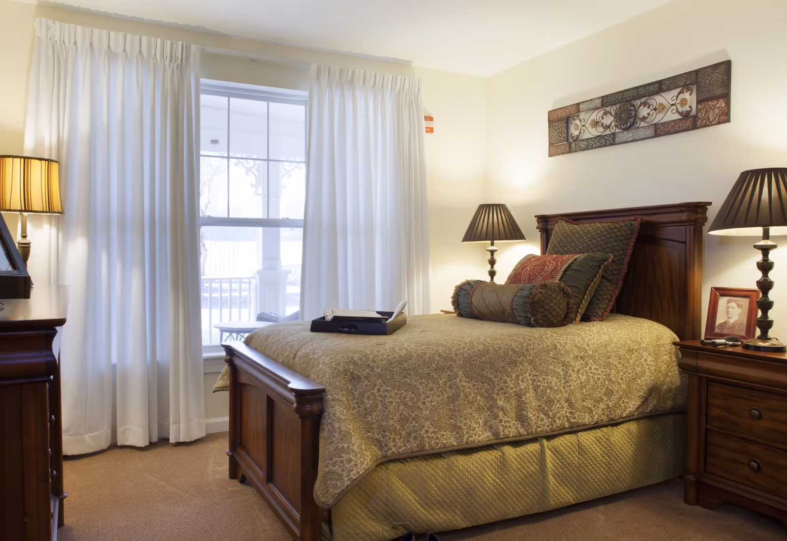 A cozy bedroom with a wooden bed frame and a beige patterned bedspread. The bed is adorned with multiple decorative pillows. There are two matching wooden nightstands on either side of the bed, each with a lamp. A framed photograph is on the right nightstand. White curtains cover a window behind the bed, and a decorative wall art piece hangs above the headboard.