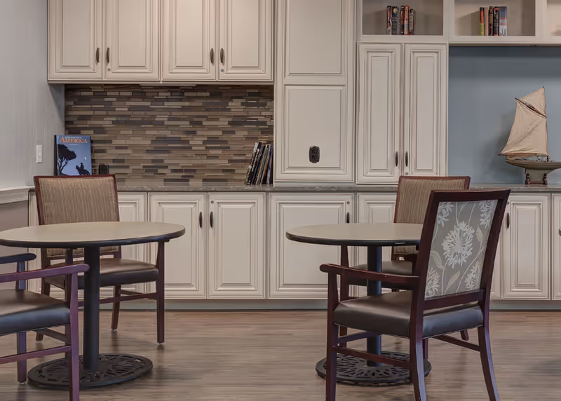A room with two round tables and four chairs with wooden frames and cushioned seats. Behind the tables is a wall with white cabinetry and a decorative tiled backsplash. On the countertop are a few books and a small model sailboat. The floor is wooden.