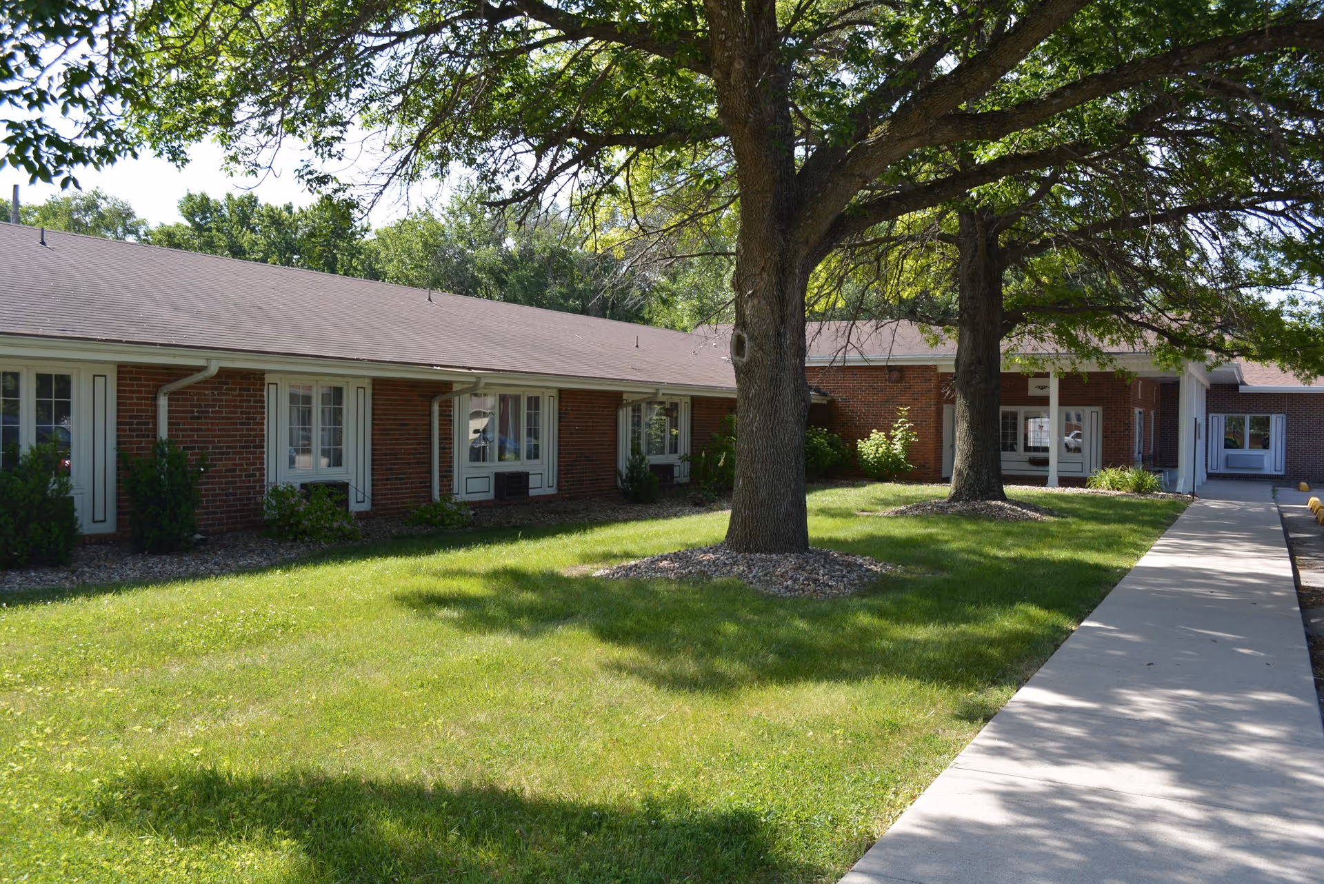 Exterior view of a single-story brick building with multiple windows and a sidewalk leading to an entrance. The building is surrounded by green grass, trees providing shade, and some small bushes near the windows.