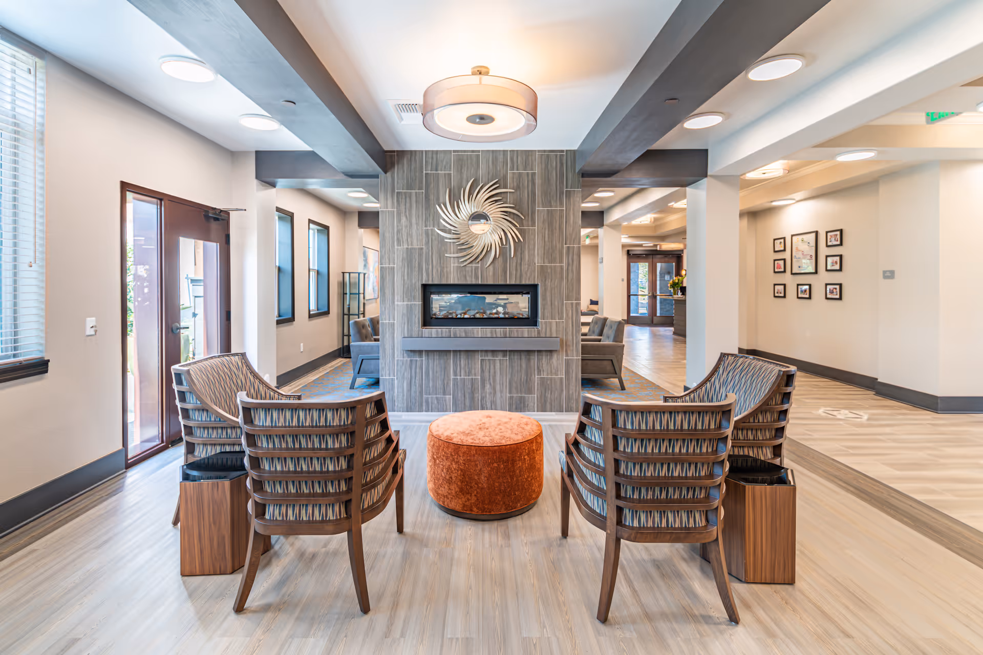 A bright and modern senior living facility common area with four wooden chairs arranged around a round orange ottoman in front of a decorative wall with a built-in electric fireplace and a sunburst mirror above it. The room has light wood flooring, large windows with blinds, and recessed ceiling lights. There are additional seating areas and framed pictures on the walls in the background.