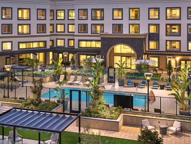 Evening view of an outdoor courtyard at Cogir of South Bay featuring a swimming pool surrounded by lounge chairs, potted plants, and landscaped garden areas with a multi-story building in the background with lit windows.