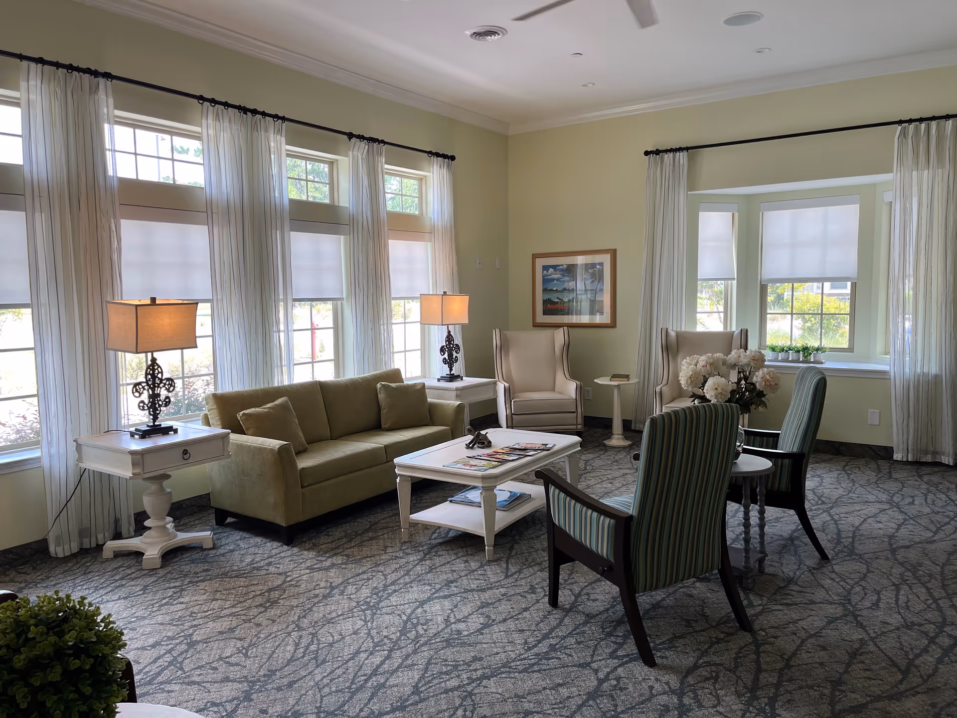 Sunlit sitting area with a sofa, armchairs, coffee table, lamps, and large windows with sheer curtains.