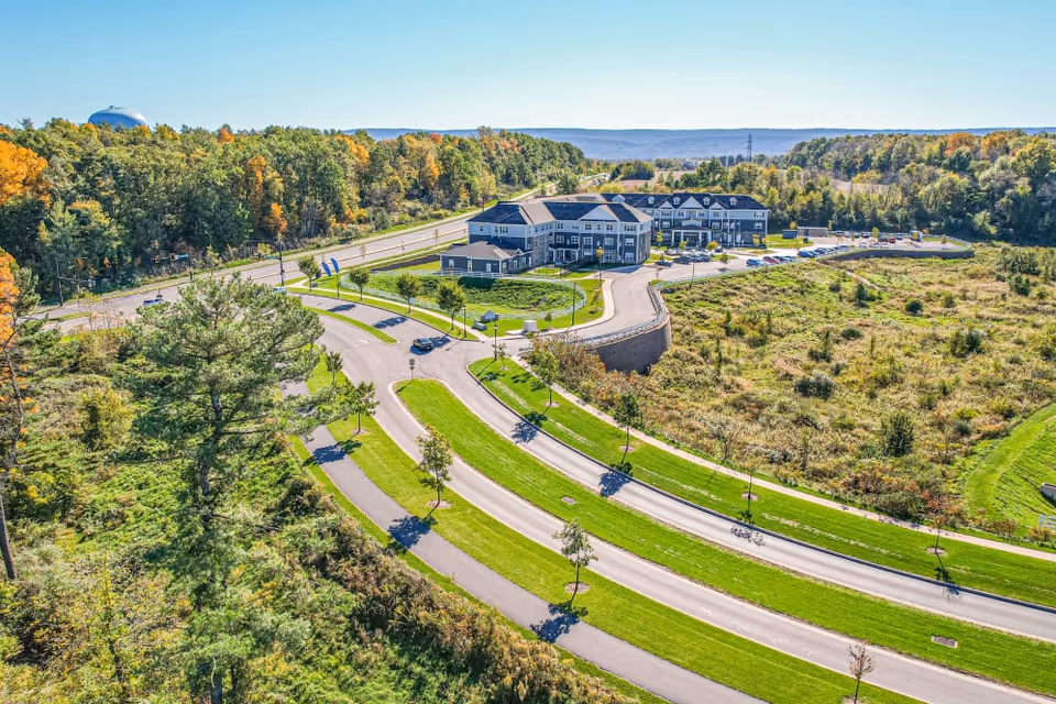Aerial view of Harmony at State College facility surrounded by greenery and trees with a curved road leading to the building. The building is a multi-story structure with a parking lot and is set against a backdrop of forested hills under a clear blue sky.