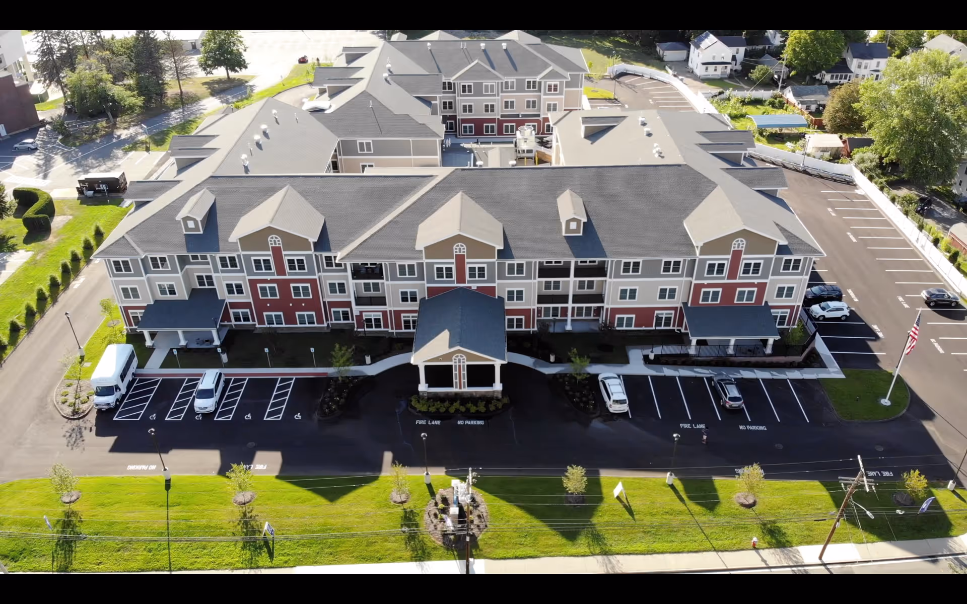 Aerial view of a large, multi-story senior living facility building with a gray roof and beige and red exterior walls. The building has multiple windows and covered entrances. There are several parking spaces, including handicapped spots, and landscaped green areas surrounding the facility. The facility is situated in a suburban neighborhood with other houses and trees visible in the background.