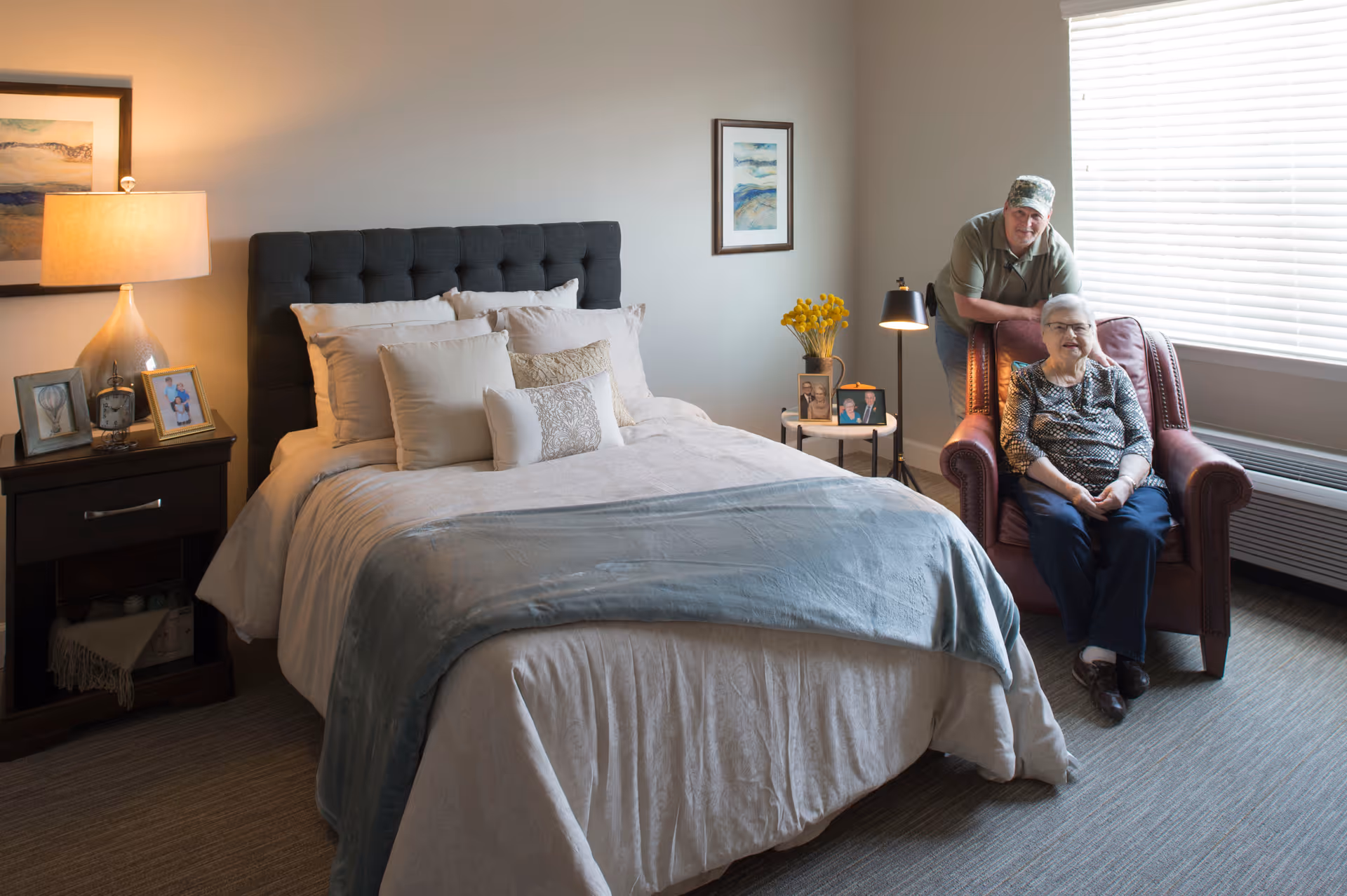 A cozy bedroom in an assisted living facility with a neatly made bed featuring multiple pillows and a blue throw blanket. To the right, an elderly woman sits in a red armchair near a window with blinds, while a man stands behind her smiling. The room is softly lit by a table lamp on a nightstand with framed photos and a clock, and a small side table with yellow flowers and more framed photos.