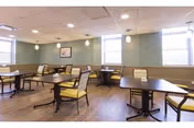 A dining room with multiple square tables and cushioned chairs arranged neatly. The room has light green walls, large windows letting in natural light, and a wooden floor. There is a framed picture on the wall and ceiling lights providing additional illumination.