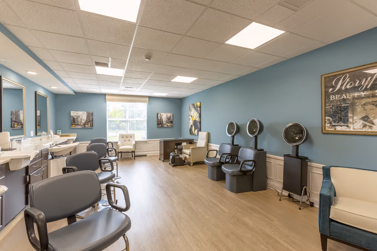 A bright and spacious beauty salon room with blue walls and wood flooring. The room features multiple salon chairs, hair dryers, sinks with mirrors, and comfortable seating. There are framed pictures on the walls and a large window letting in natural light.