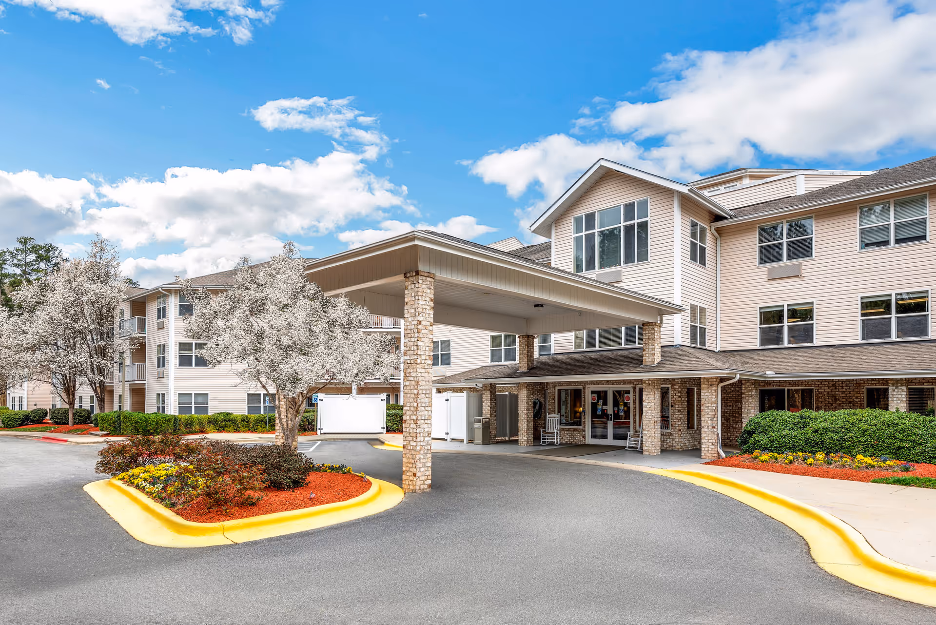 Exterior view of Solista Durham senior living facility showing a covered entrance with brick pillars, landscaped flower beds, and blooming trees under a partly cloudy blue sky.