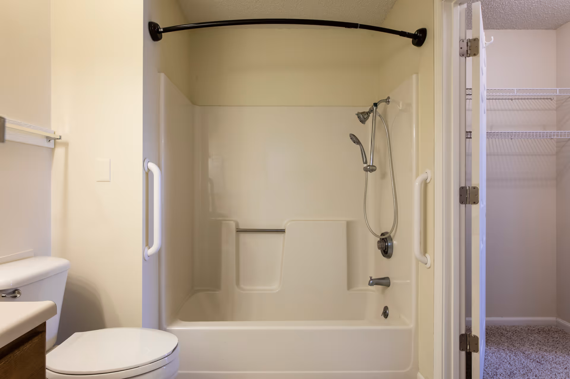 A bathroom featuring a white bathtub with built-in seating and grab bars on both sides. There is a showerhead and a handheld shower attached to the wall. To the left, a white toilet is partially visible, and to the right, an open door reveals a closet with wire shelving.