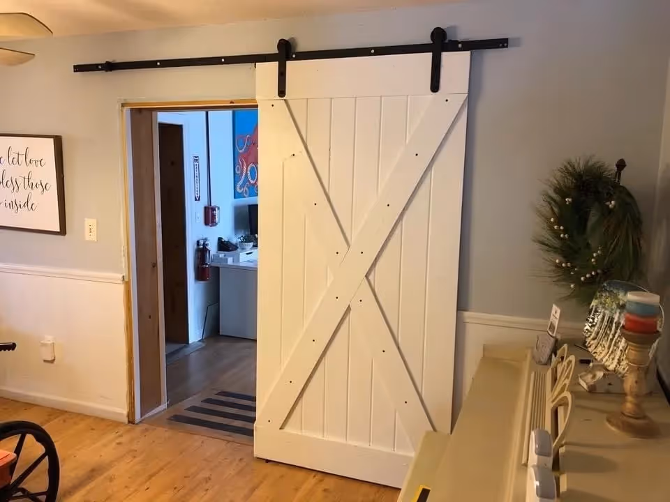 Interior view of a room with light wood flooring and light gray walls. A white sliding barn door with black hardware is partially open, revealing another room with a desk, fire extinguisher, and colorful wall art. To the right of the door is a small table or shelf decorated with a green wreath, candles, and other small decorative items. On the left wall, there is a framed sign with cursive writing.