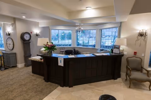 Reception area with a dark wooden front desk, two table lamps, a bouquet of flowers, and a large window showing an outdoor view. The room has beige walls, carpet and tile flooring, a grandfather clock, wall sconces, and a vintage-style chair.