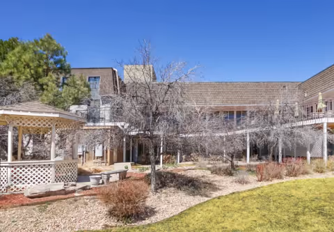 Outdoor courtyard area of a senior living facility with a gazebo, leafless trees, shrubs, and a two-story building surrounding the courtyard under a clear blue sky.