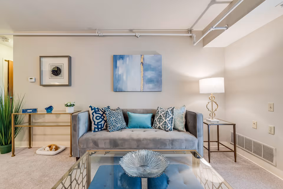 A stylish living room with a gray sofa adorned with patterned throw pillows, a glass coffee table, side tables and a lamp beneath blue wall art.