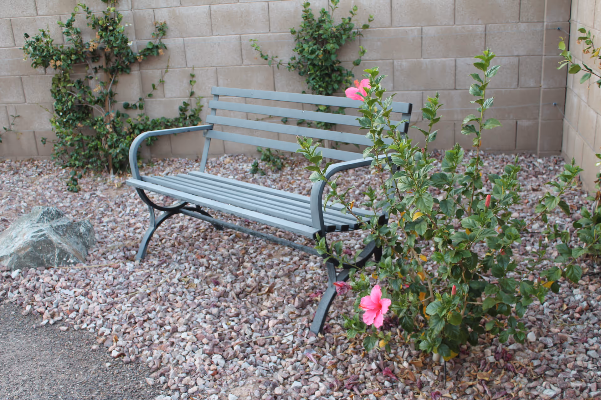 A gray metal bench placed on a bed of small rocks in an outdoor garden area. There are green plants with pink flowers growing nearby, and a beige brick wall in the background.