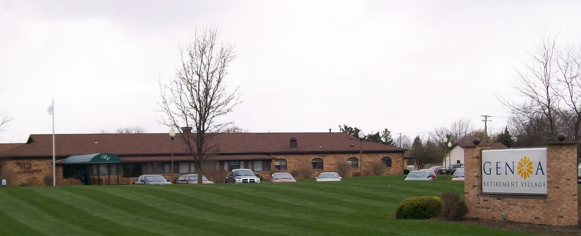 Exterior view of Genoa Retirement Village building with a well-maintained lawn, several parked cars, leafless trees, and a large sign displaying the facility name.
