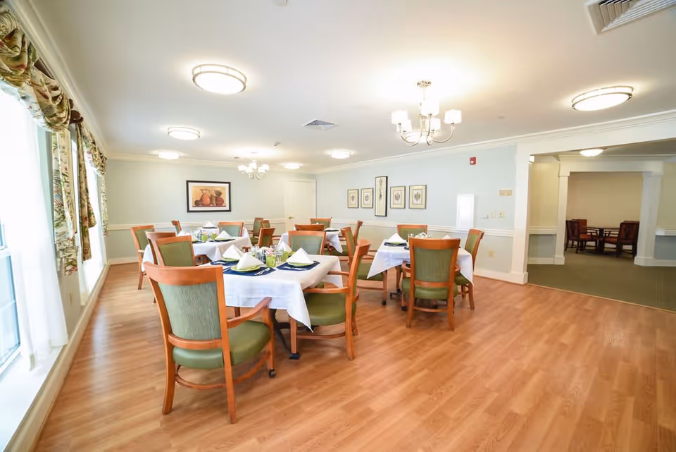 A bright dining room with wooden floors and several tables set with white tablecloths, napkins, and place settings. The room has green cushioned wooden chairs, framed artwork on the walls, and large windows with floral curtains letting in natural light. Ceiling lights and a chandelier illuminate the space.