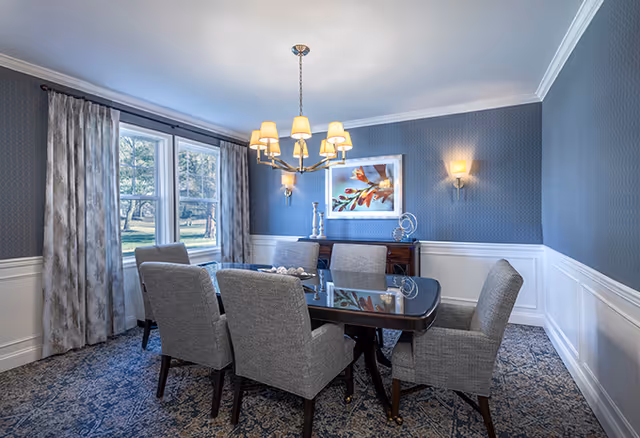 A dining room with a rectangular dark wooden table surrounded by six gray upholstered chairs. The room has blue patterned wallpaper with white wainscoting, a large window with gray curtains, a chandelier with multiple lampshades hanging above the table, and a framed artwork on the wall. There is also a sideboard with decorative items beneath the artwork.