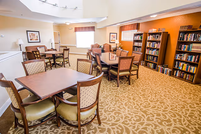 A well-lit common area with multiple tables and chairs arranged for seating. The room features patterned carpet flooring, bookshelves filled with books along one wall, framed artwork, and windows with blinds allowing natural light to enter.