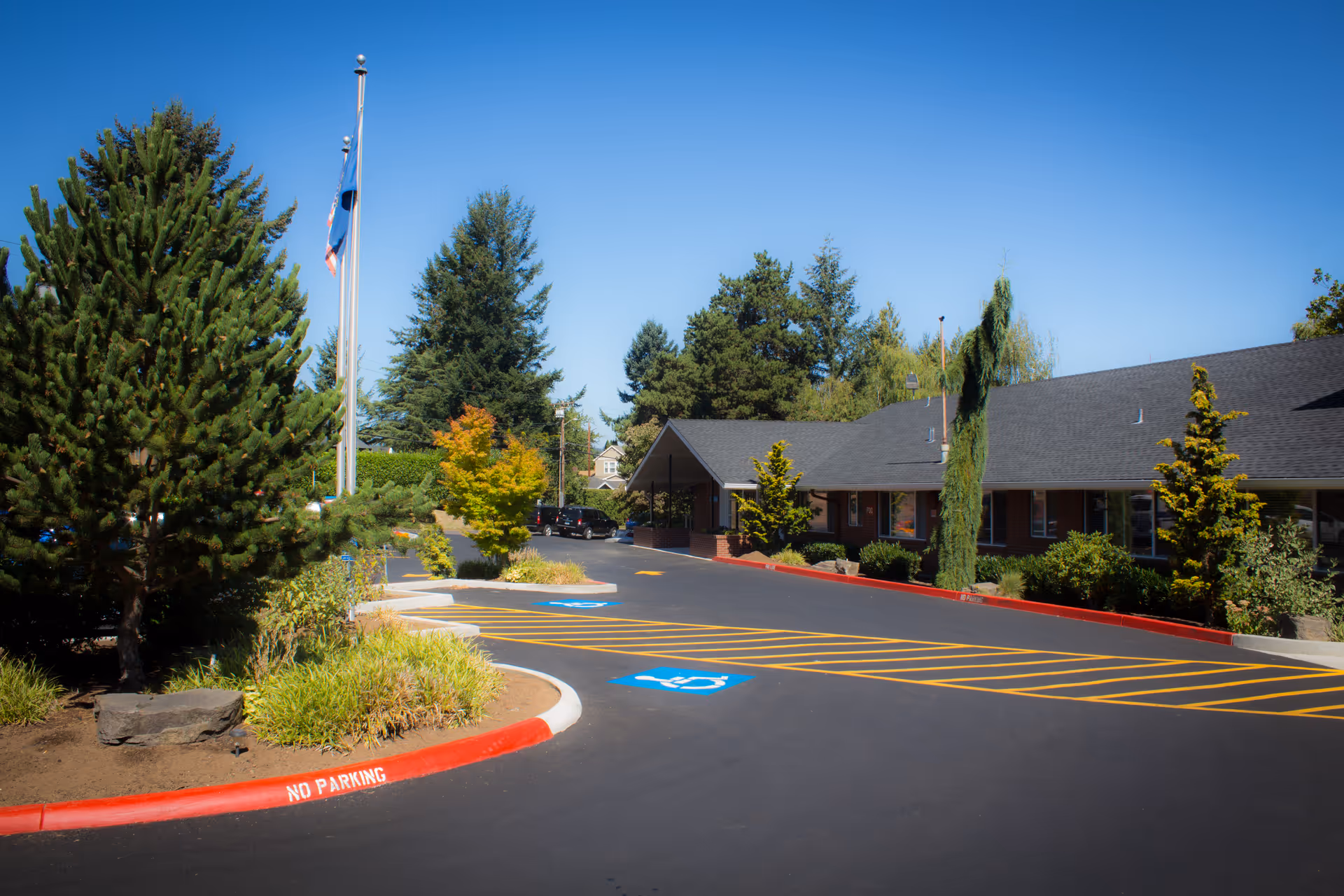 Exterior view of Avamere Crestview of Portland showing a paved parking area with handicap parking spaces and a no parking zone marked in red. The building is single-story with a dark roof and surrounded by trees and shrubs under a clear blue sky.