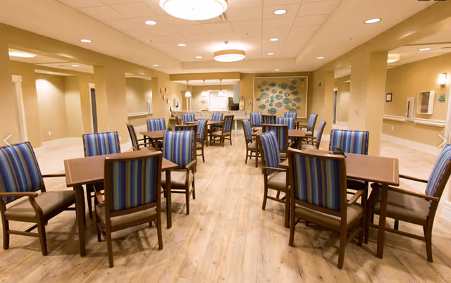A spacious dining room with multiple wooden tables and chairs featuring blue and green striped upholstery. The room has light-colored wooden flooring, beige walls, and a ceiling with recessed lighting and a central round light fixture. In the background, there is a decorative wall with a fish-themed art installation and a kitchen area with white cabinets and appliances.