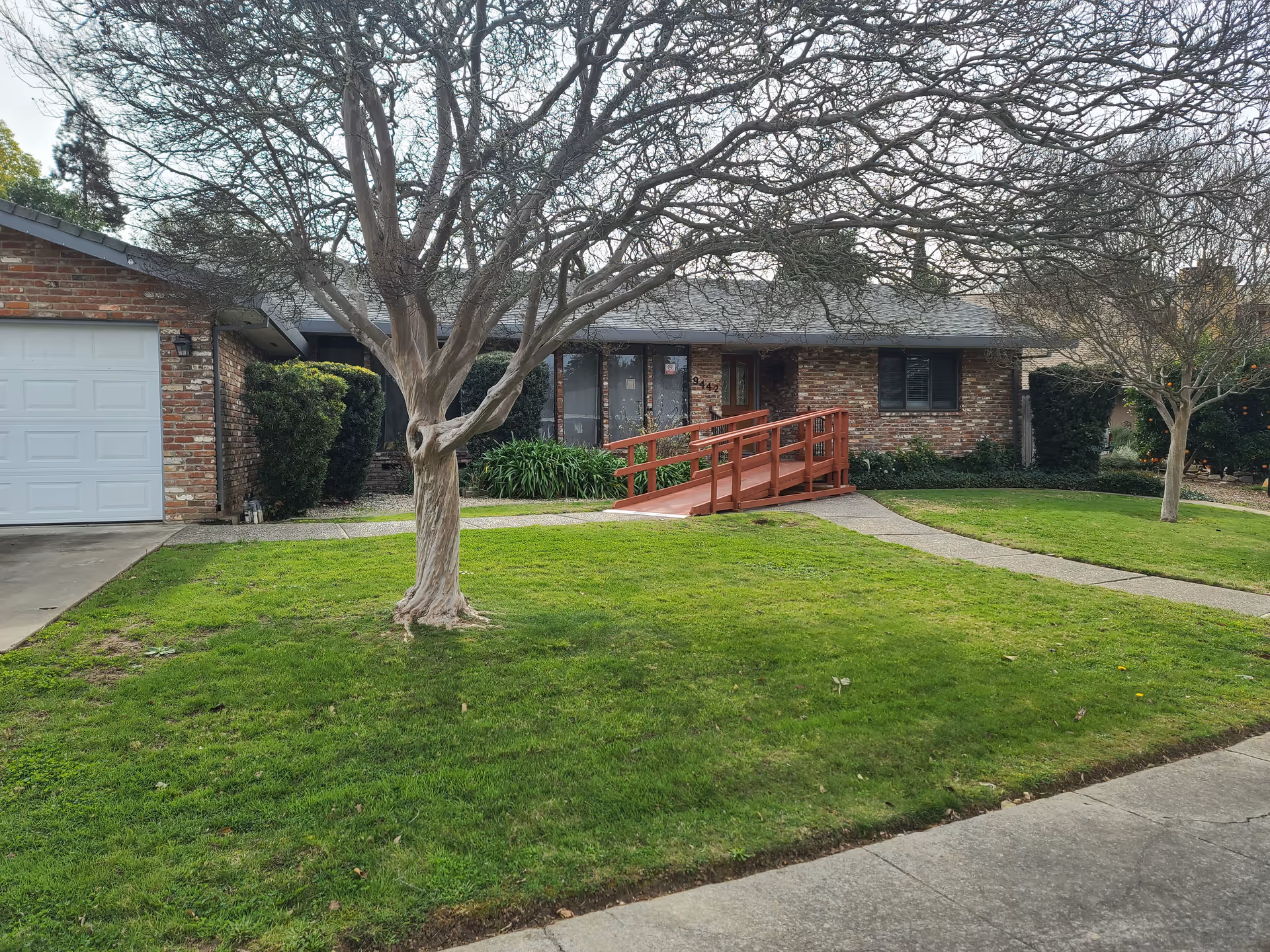 Brick single-story house with a red wooden wheelchair ramp, front lawn and leafless trees.