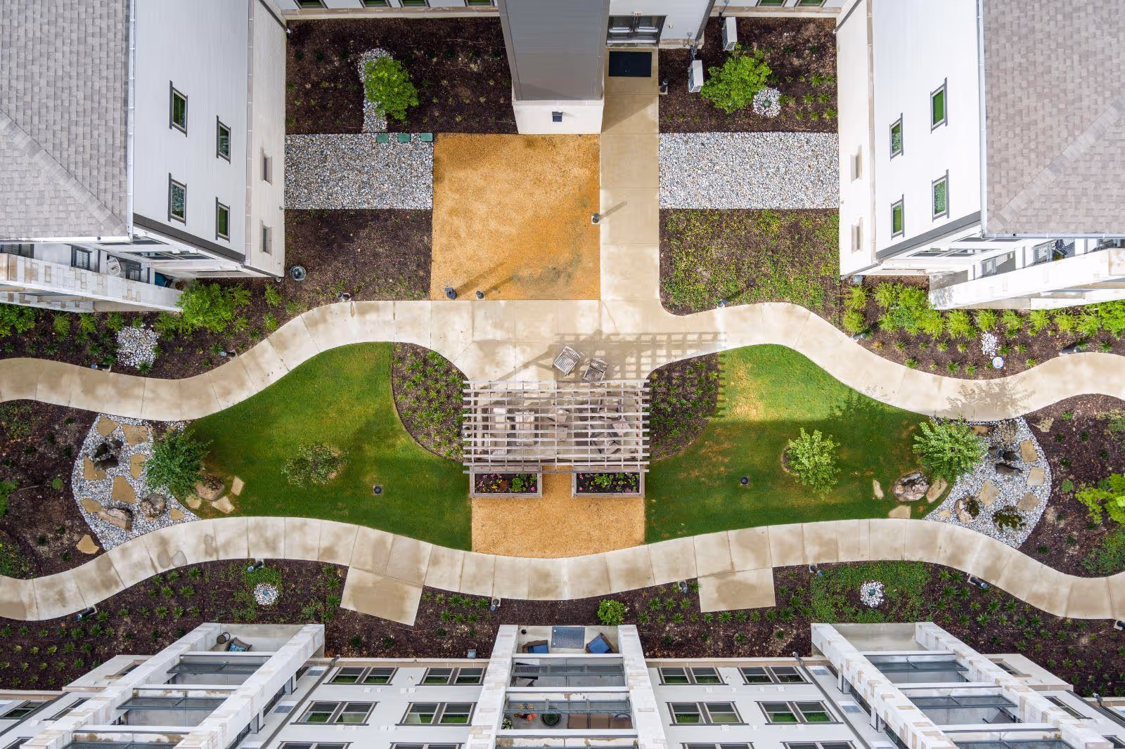 Aerial view of a landscaped courtyard between residential buildings at Hidden Springs of McKinney, featuring winding concrete pathways, green grass areas, small trees, decorative rocks, and a wooden pergola with seating underneath.