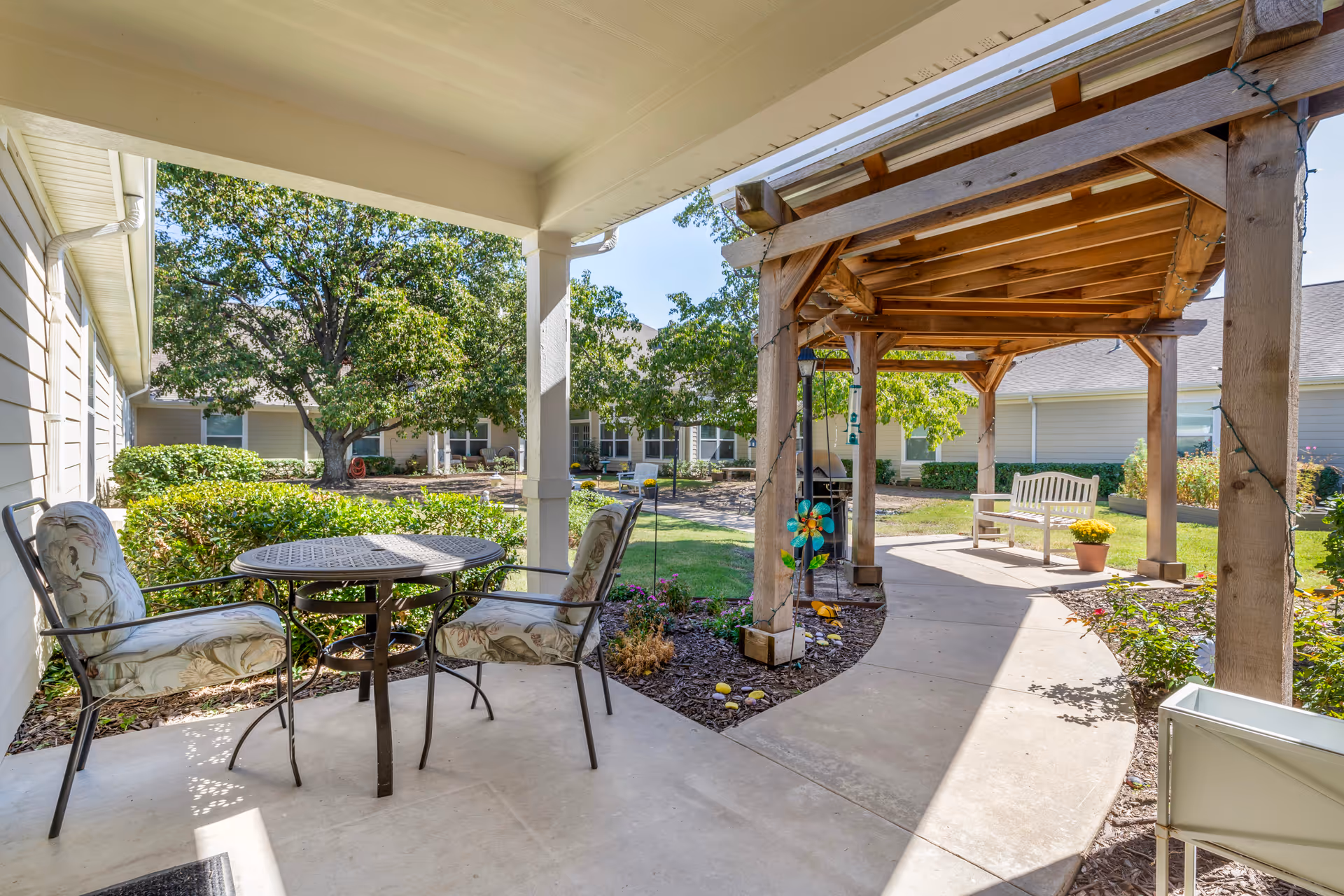 Outdoor patio area at Brookdale Owasso featuring a small round metal table with two cushioned chairs under a covered porch. A wooden pergola with string lights and a bench is visible along a curved concrete pathway surrounded by green bushes, trees, and a well-maintained garden.