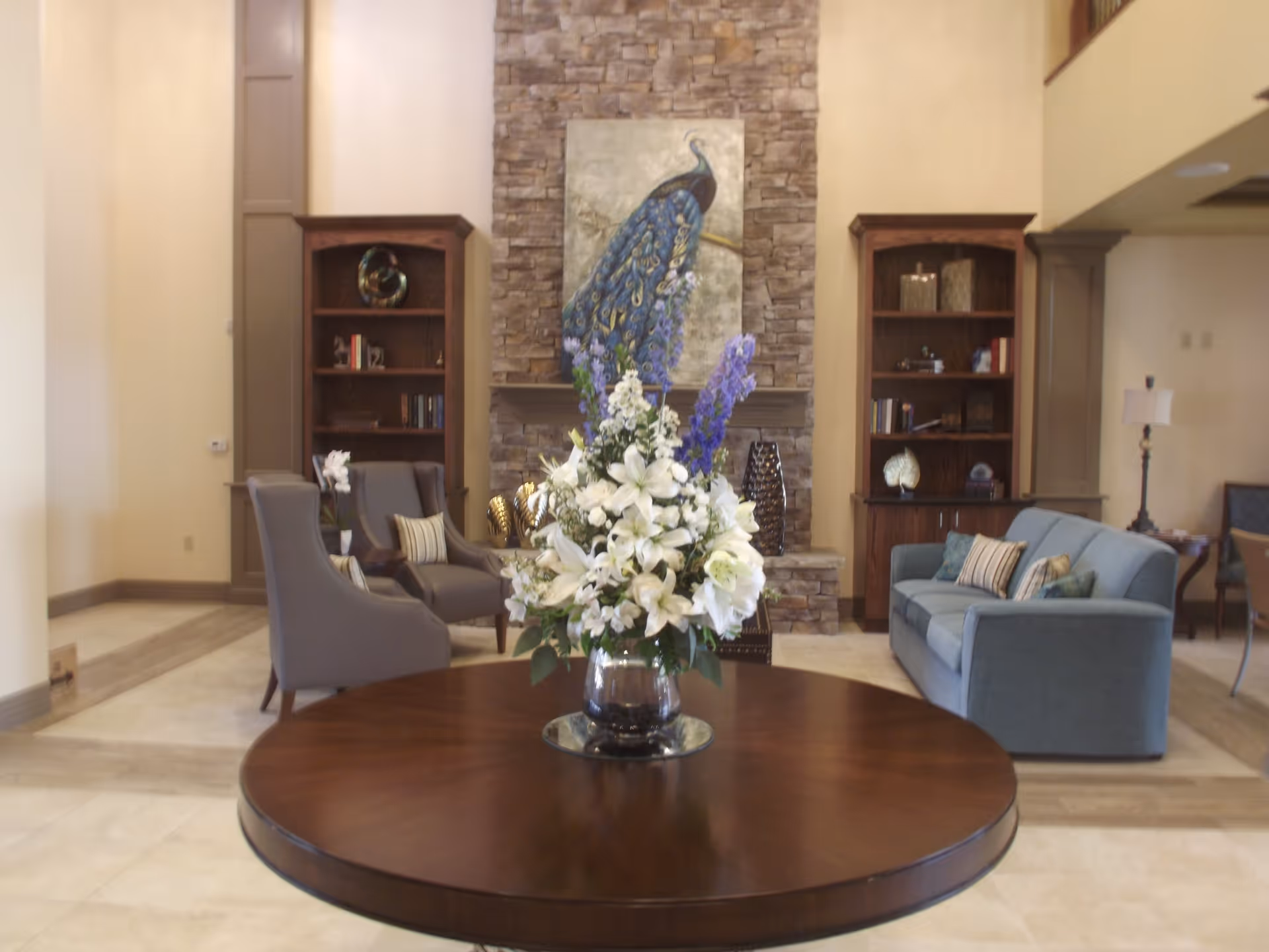 A cozy living room area with a round wooden table in the foreground holding a large bouquet of white and purple flowers. Behind the table, there are two gray armchairs with striped pillows on the left, a blue sofa with pillows on the right, and two wooden bookshelves flanking a stone fireplace with a peacock painting above it.