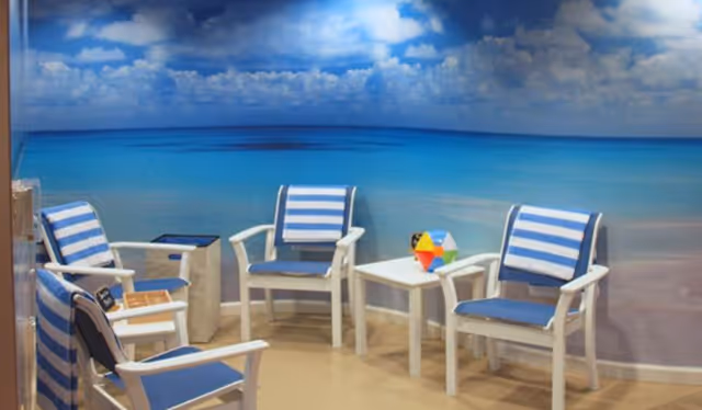 A small seating area with four white chairs featuring blue cushions and blue-and-white striped towels draped over the backs. A white table is placed in the center with a colorful geometric toy on it. The background is a large wall mural depicting a serene beach scene with blue sky, clouds, and calm ocean water.
