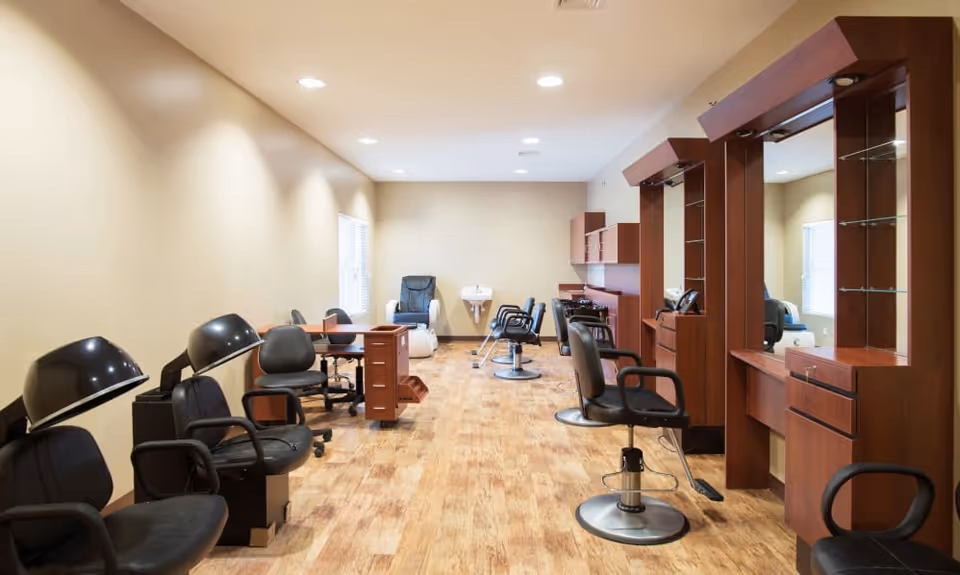 Interior view of a salon area with multiple black salon chairs, hair drying stations on the left, and wooden styling stations with mirrors on the right. The room has light-colored walls, wooden flooring, and recessed ceiling lights.