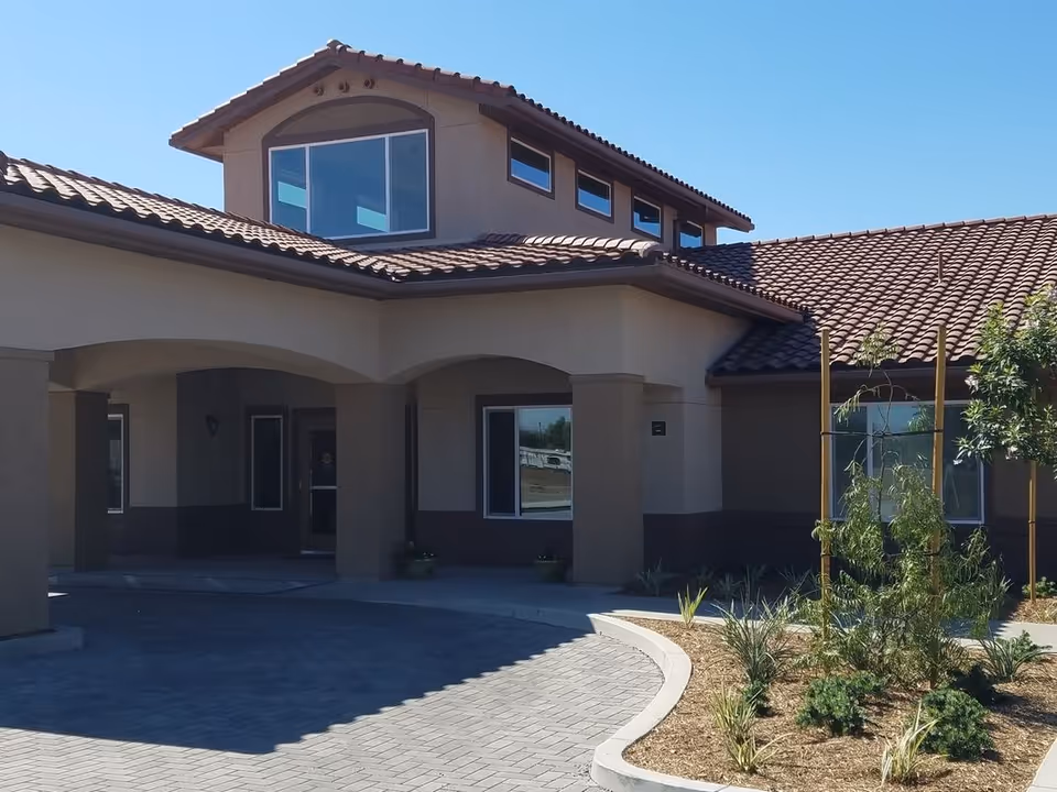 Front entrance of a building with a covered porte-cochère, tiled roof, and landscaped driveway.