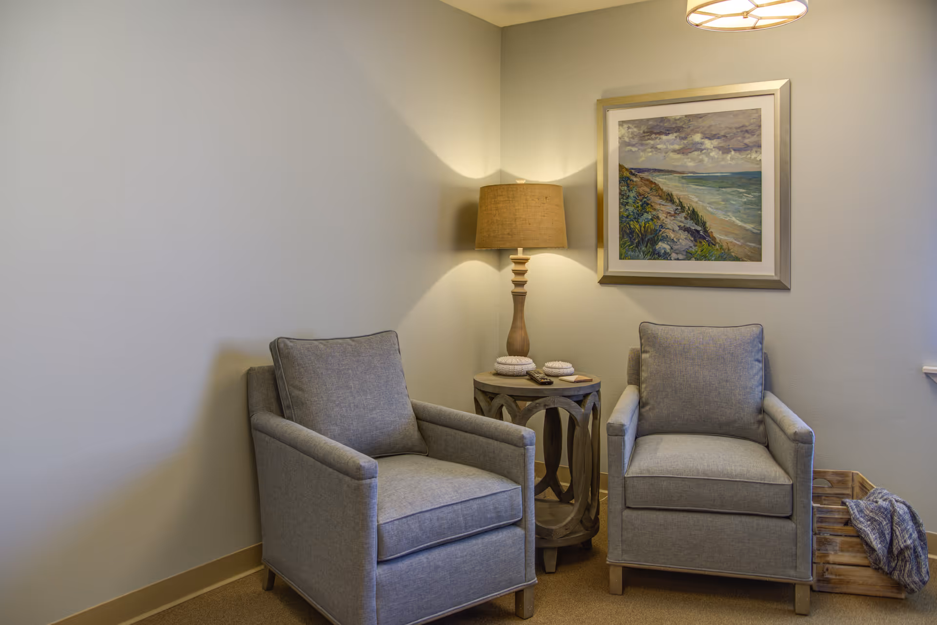 A cozy corner with two gray upholstered armchairs separated by a small round wooden side table holding a lamp and decorative items. Above the table hangs a framed painting of a coastal landscape. A wooden crate with a blanket is placed on the floor to the right.