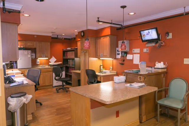 Interior view of a healthcare facility office area with wooden cabinets, desks, office chairs, a countertop with paperwork, a water pitcher, and a wall-mounted TV. The walls are painted orange and the floor is wooden.