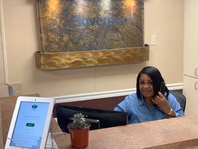 Reception area at Clovernook Health Care LLC with a woman sitting behind the front desk talking on the phone. A computer monitor and a small potted succulent plant are on the desk. A sign with the Clovernook logo is mounted on the wall behind her.