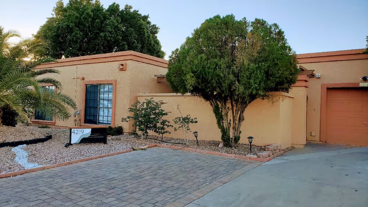 Exterior view of a single-story building with beige walls and orange trim, featuring a large window with blinds, a garage door, and a driveway made of stone pavers. There are various green trees and shrubs around the building, along with a landscaped area with rocks and a small sign displaying the number 4219.