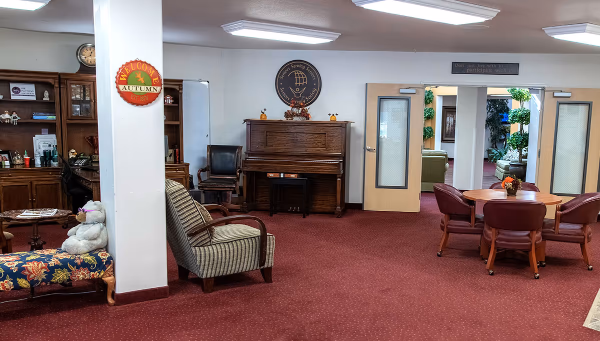 Interior of a senior living facility lounge area with red carpet, a wooden piano against the wall, a round table with four red chairs, a striped armchair, a floral bench with a teddy bear wearing purple glasses, and wooden cabinets. There are double doors leading to another room with plants and seating visible.
