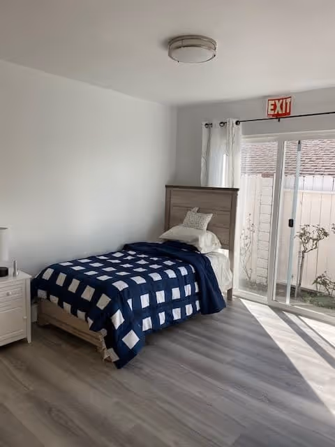 Minimal bedroom with a twin bed dressed in a navy-and-white quilt, a nightstand, sliding glass door with an EXIT sign, and light wood-look flooring.
