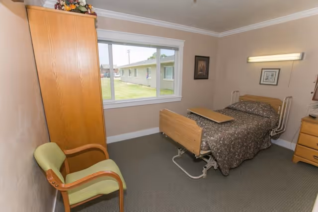 Small furnished bedroom with an adjustable single bed, bedside furniture, a chair, and a window overlooking a lawn.