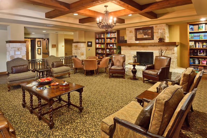 A cozy living room area in an assisted living facility featuring patterned carpet, multiple upholstered chairs and sofas, a wooden coffee table with decorative items, a stone fireplace with a painting above it, built-in bookshelves filled with books, and a chandelier hanging from a wooden beam ceiling.